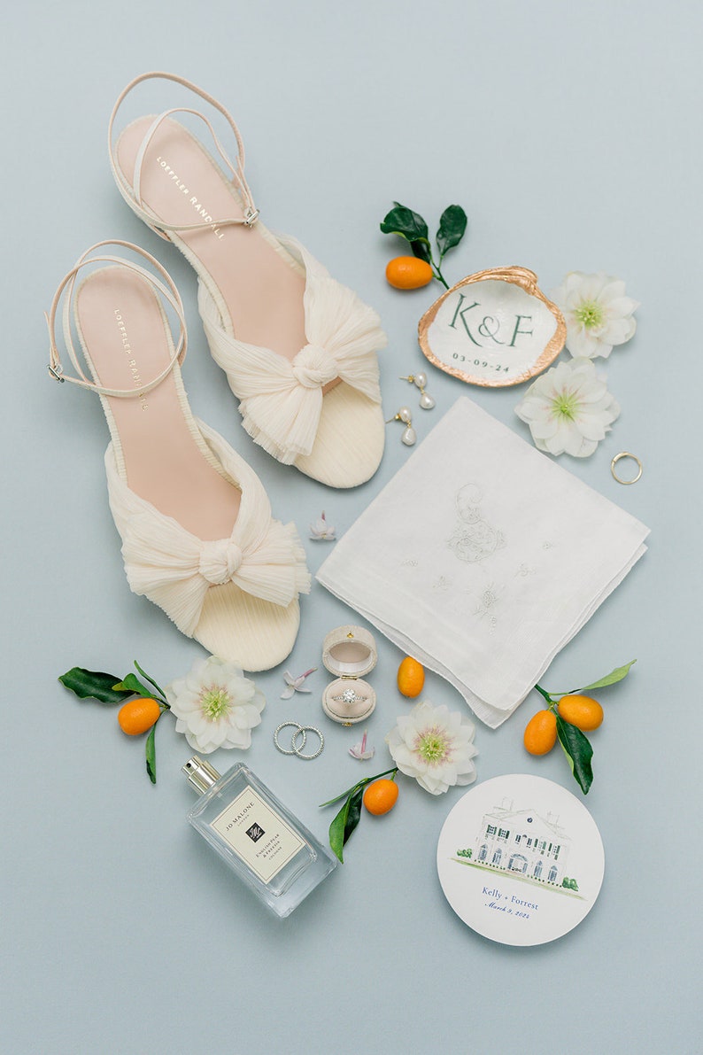 May include: A pair of cream-colored pleated sandals with ankle straps, a white embroidered handkerchief, a perfume bottle, a ring in a velvet box, two wedding bands, a painted shell with K&F 03-09-24, a round card with a house illustration, and kumquats.
