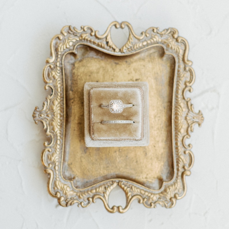 May include: Two gold rings with diamonds are displayed in a small, beige velvet box. The box is sitting on a gold, ornate tray.
