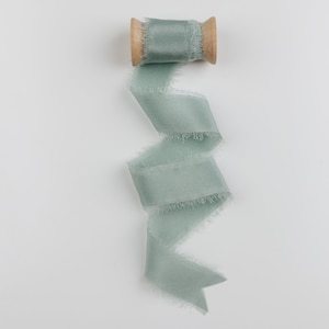 May include: A spool of sage green ribbon with frayed edges. The ribbon is wound around a wooden spool.
