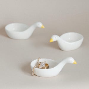 May include: Three white ceramic goose-shaped ring dishes with yellow beaks. One dish holds a gold ring and a diamond ring, and a gold chain. The other two dishes are empty. The dishes are on a neutral background.