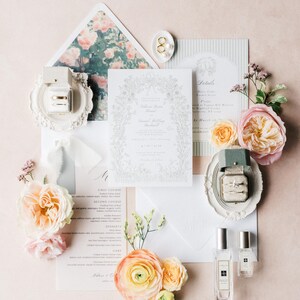 Puede incluir: Un conjunto de invitaciones de boda con arreglos florales, papeler&iacute;a y frascos de perfume. La tarjeta de invitaci&oacute;n tiene un borde floral y los nombres "Rebecca Ryan" y "Daniel Willoughby" son visibles. Anillos de oro se exhiben en un plato decorativo blanco.