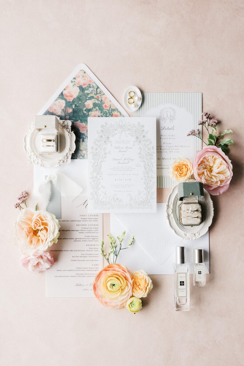 Puede incluir: Un conjunto de invitaciones de boda con arreglos florales, papeler&iacute;a y frascos de perfume. La tarjeta de invitaci&oacute;n tiene un borde floral y los nombres "Rebecca Ryan" y "Daniel Willoughby" son visibles. Anillos de oro se exhiben en un plato decorativo blanco.