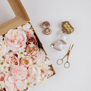 May include: A craft box filled with pink and cream-colored flowers, a ring in a pink velvet box, a gold thimble, a measuring tape, and gold scissors. The items are arranged on a white surface.
