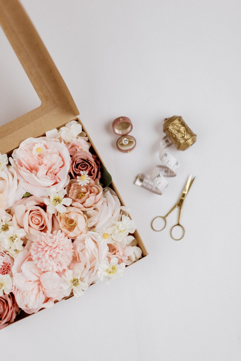May include: A craft box filled with pink and cream-colored flowers, a ring in a pink velvet box, a gold thimble, a measuring tape, and gold scissors. The items are arranged on a white surface.