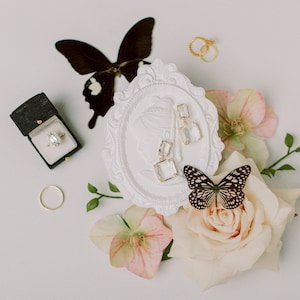 Puede incluir: Una elegante composici&oacute;n con un anillo de compromiso de diamantes en una caja negra, anillos de oro y pendientes cuadrados transparentes. Mariposas y flores, incluyendo rosas y hel&eacute;boros, a&ntilde;aden un toque delicado. Fondo neutro.