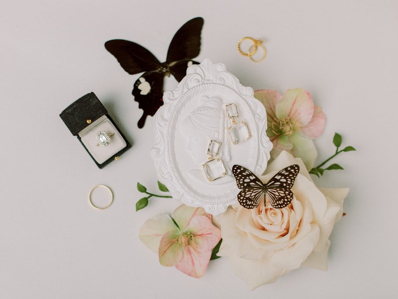 Puede incluir: Una elegante composici&oacute;n con un anillo de compromiso de diamantes en una caja negra, anillos de oro y pendientes cuadrados transparentes. Mariposas y flores, incluyendo rosas y hel&eacute;boros, a&ntilde;aden un toque delicado. Fondo neutro.