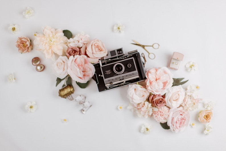 May include: A vintage black Polaroid camera is surrounded by a collection of blush and cream-colored roses and other flowers. Gold scissors, a ring box, and earrings are also arranged on a white surface, creating a romantic, floral theme.