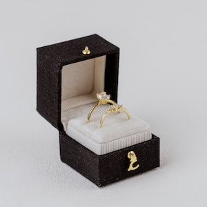 Puede incluir: Dos anillos de oro con piedras preciosas transparentes presentados en un joyero marr&oacute;n oscuro abierto con un cierre dorado. El joyero tiene un interior beige claro y un emblema dorado en la parte delantera. Los anillos est&aacute;n sobre una superficie blanca y aterciopelada.