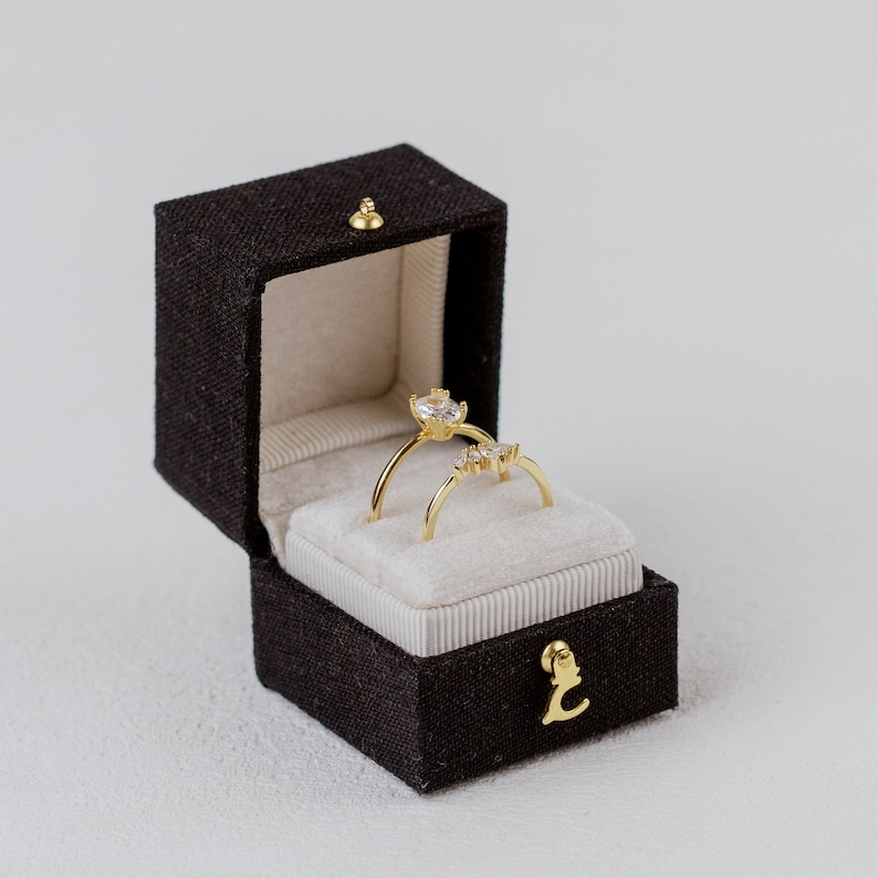 Puede incluir: Dos anillos de oro con piedras preciosas transparentes presentados en un joyero marr&oacute;n oscuro abierto con un cierre dorado. El joyero tiene un interior beige claro y un emblema dorado en la parte delantera. Los anillos est&aacute;n sobre una superficie blanca y aterciopelada.