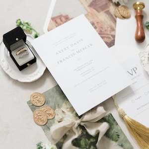 Puede incluir: Conjunto de papeler&iacute;a de boda con una invitaci&oacute;n formal con los nombres "AVERY DAVIS y FRANCIS MERCER". Incluye una caja negra para anillos con dos anillos, sellos de cera dorados y una borla dorada. El conjunto tiene una paleta de colores neutros.