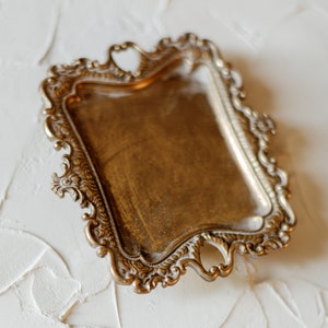 May include: A gold-toned metal tray with an ornate, decorative design. The tray has a rectangular shape with a raised edge and intricate details around the perimeter.