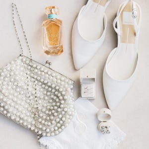 Puede incluir: Vista a&eacute;rea de accesorios de novia: zapatos blancos con correa trasera, un bolso de mano cubierto de perlas, un frasco de perfume y un pa&ntilde;uelo con iniciales bordadas. Tambi&eacute;n se ven joyeros y un rosario con cruz.