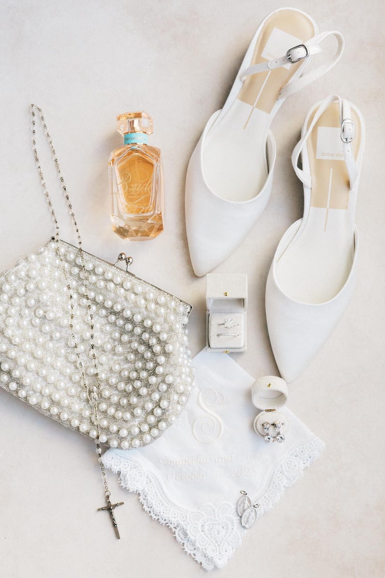 Puede incluir: Vista a&eacute;rea de accesorios de novia: zapatos blancos con correa trasera, un bolso de mano cubierto de perlas, un frasco de perfume y un pa&ntilde;uelo con iniciales bordadas. Tambi&eacute;n se ven joyeros y un rosario con cruz.