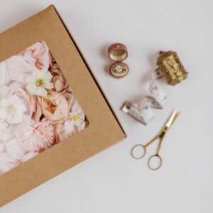 May include: A brown cardboard box with a clear window filled with pink and white flowers. A ring in a pink velvet box, a gold thimble, a measuring tape, and gold scissors are also present.