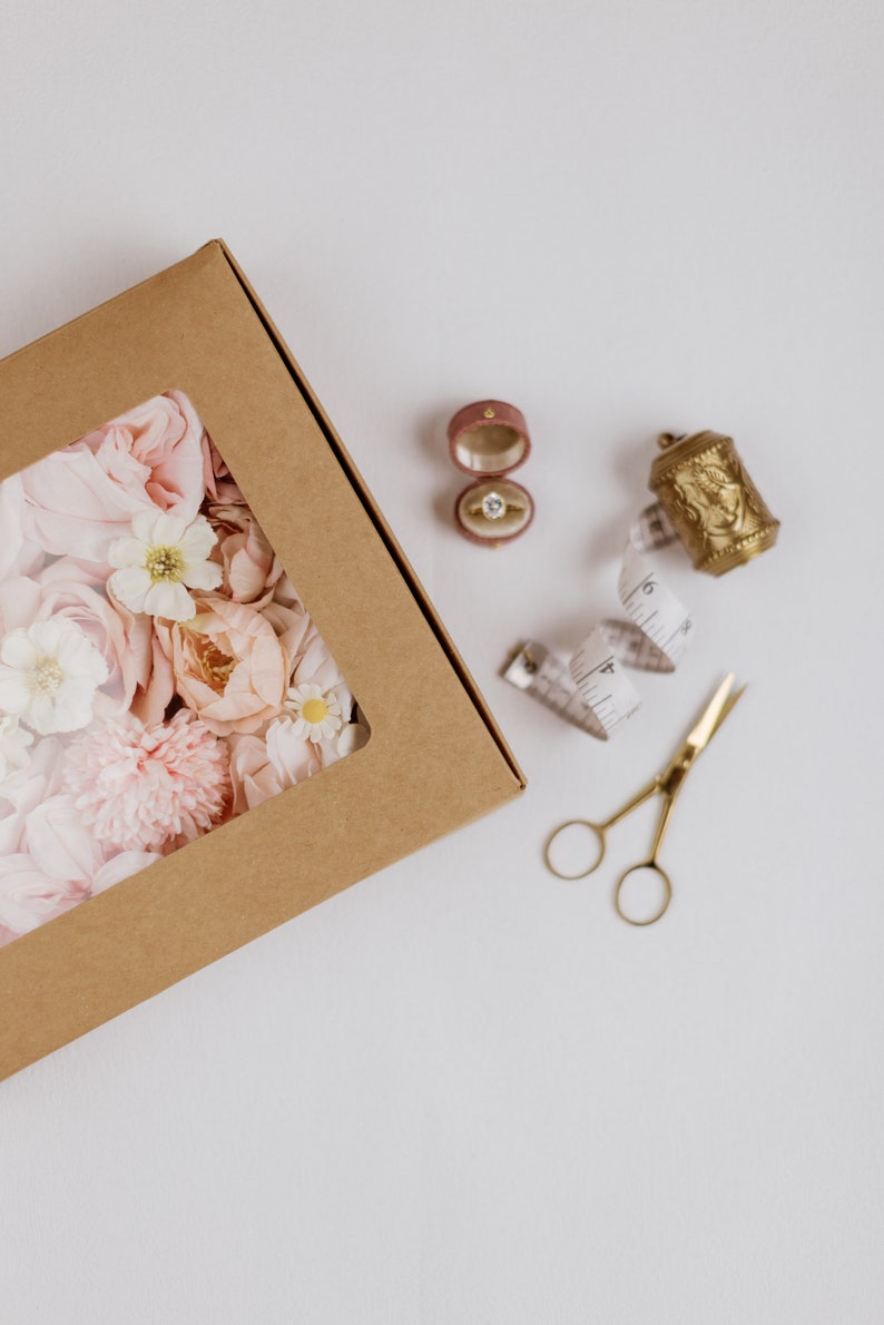 May include: A brown cardboard box with a clear window filled with pink and white flowers. A ring in a pink velvet box, a gold thimble, a measuring tape, and gold scissors are also present.