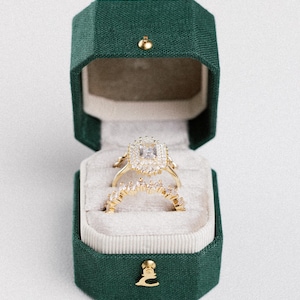May include: A gold engagement ring set with a large, square-cut diamond center stone and smaller surrounding stones. The ring sits in an open, emerald green velvet ring box with a gold clasp. The set includes a matching band.