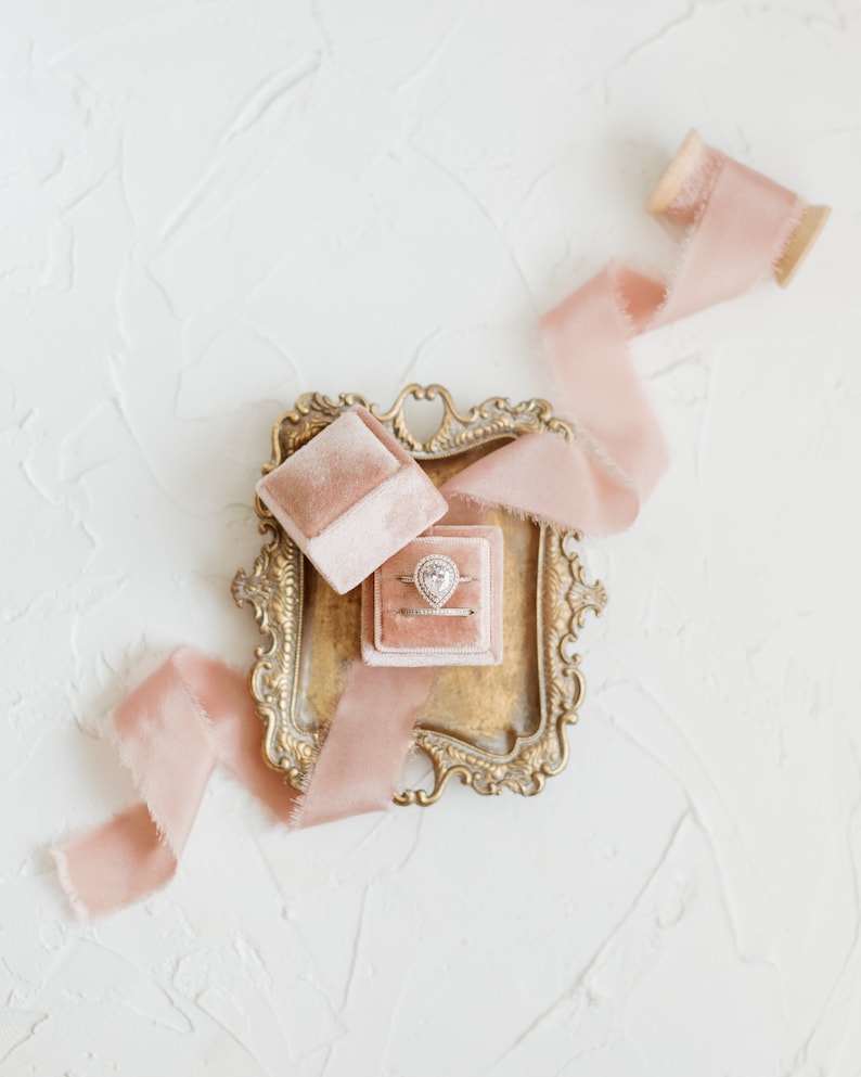 May include: A pear-shaped engagement ring with a diamond center stone in a pink velvet ring box. The ring box is on a gold-colored tray with a pink ribbon.