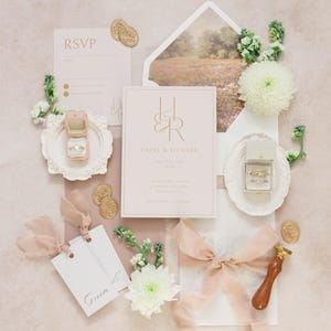 Puede incluir: Un conjunto de invitaciones de boda en tonos rosa rubor y dorado. El conjunto incluye una invitaci&oacute;n, una tarjeta de confirmaci&oacute;n de asistencia y etiquetas, junto con un joyero, sellos de cera y detalles florales. La invitaci&oacute;n muestra los nombres "Hazel & Richard".