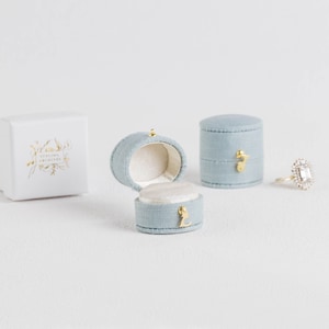 May include: Two oval light blue fabric ring boxes with gold-toned latches, one open and one closed, alongside a white box with gold floral branding that reads STYLING ARCHIVES and a gold ring featuring a rectangular center stone and halo setting.