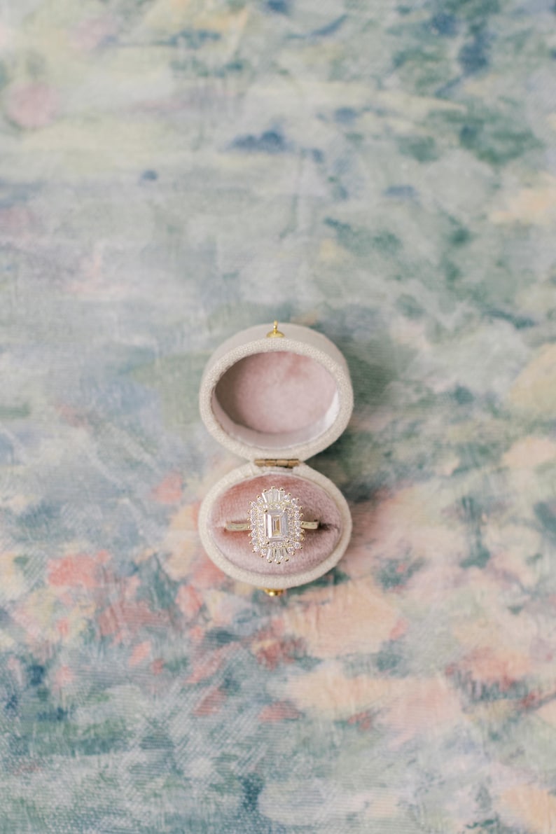 May include: An emerald-cut diamond ring with a halo of smaller stones set on a thin gold band, displayed inside an open, oval-shaped, cream-colored velvet ring box with a soft pink interior lining, showcasing a clear and elegant design.