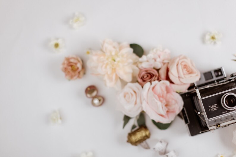 May include: A flat lay featuring a vintage Polaroid camera, blush and peach-colored roses, and small white flowers. Gold-toned earrings and a decorative gold object are also present, creating a romantic and artistic composition.