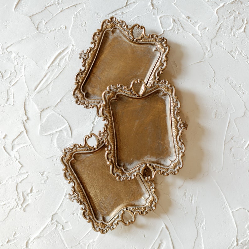 May include: Three gold-toned metal trays with ornate, decorative edges. The trays are stacked on top of each other, with the largest tray on the bottom and the smallest tray on top.
