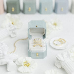 May include: A gold engagement ring and wedding band set in a light blue velvet ring box. The ring box is open, revealing the rings. Additional ring boxes and white flowers are in the background. A gold chain and a small white plate with a gold ring are also visible.
