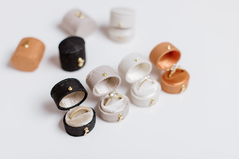 May include: A collection of small, oval-shaped velvet ring boxes in black, beige, and tan colors. Some boxes are closed, while others are open, revealing gold rings with clear stones set inside. Each box features a small gold-toned metal clasp.