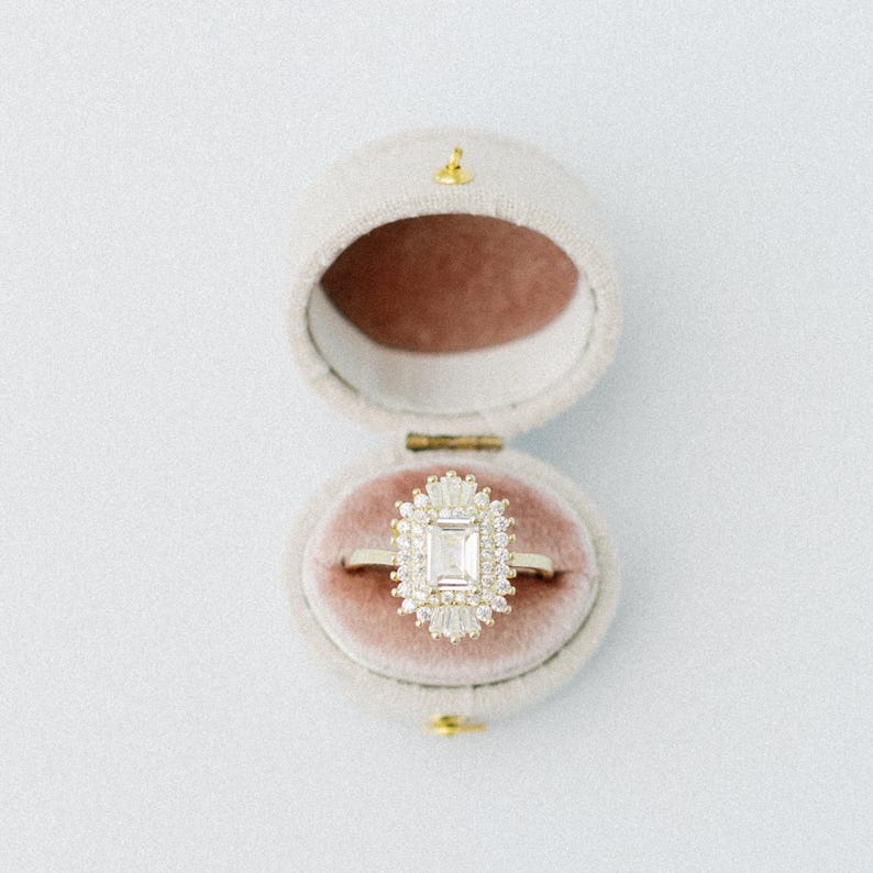 May include: A gold ring featuring a central emerald-cut stone surrounded by a halo of smaller baguette and round-cut stones, presented inside an oval-shaped, light beige velvet ring box with a dusty rose-colored interior lining.