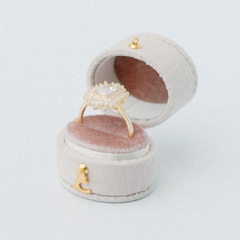 May include: A gold ring with a central oval stone surrounded by a halo of smaller clear stones, resting inside a round, beige fabric-covered ring box with a dusty pink velvet interior and a small gold-toned metal latch on the front.