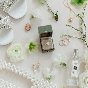 Puede incluir: Una composici&oacute;n nupcial con sandalias blancas con cristales, collar de perlas, joyero con anillo de diamantes, joyas doradas y frasco de perfume. Flores blancas y vegetaci&oacute;n se distribuyen alrededor de los accesorios.