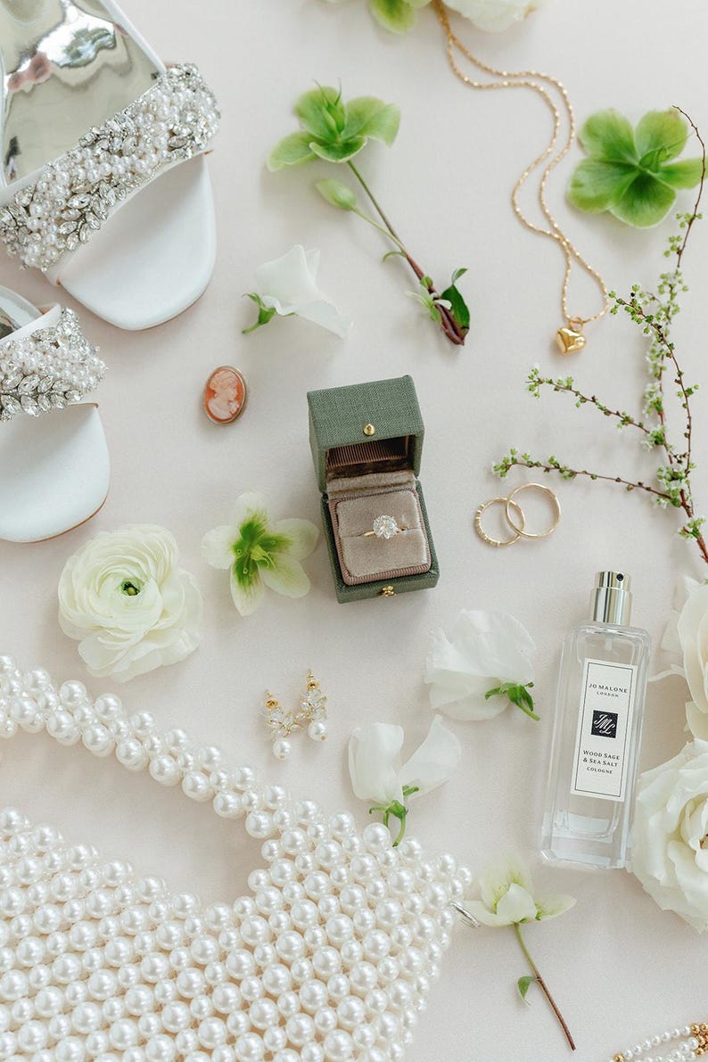 Puede incluir: Una composici&oacute;n nupcial con sandalias blancas con cristales, collar de perlas, joyero con anillo de diamantes, joyas doradas y frasco de perfume. Flores blancas y vegetaci&oacute;n se distribuyen alrededor de los accesorios.