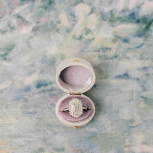 May include: A silver-toned ring featuring a central emerald-cut stone surrounded by a halo of smaller baguette and round-cut stones, resting inside a small, oval-shaped, light-colored velvet ring box with a soft, light purple interior lining.