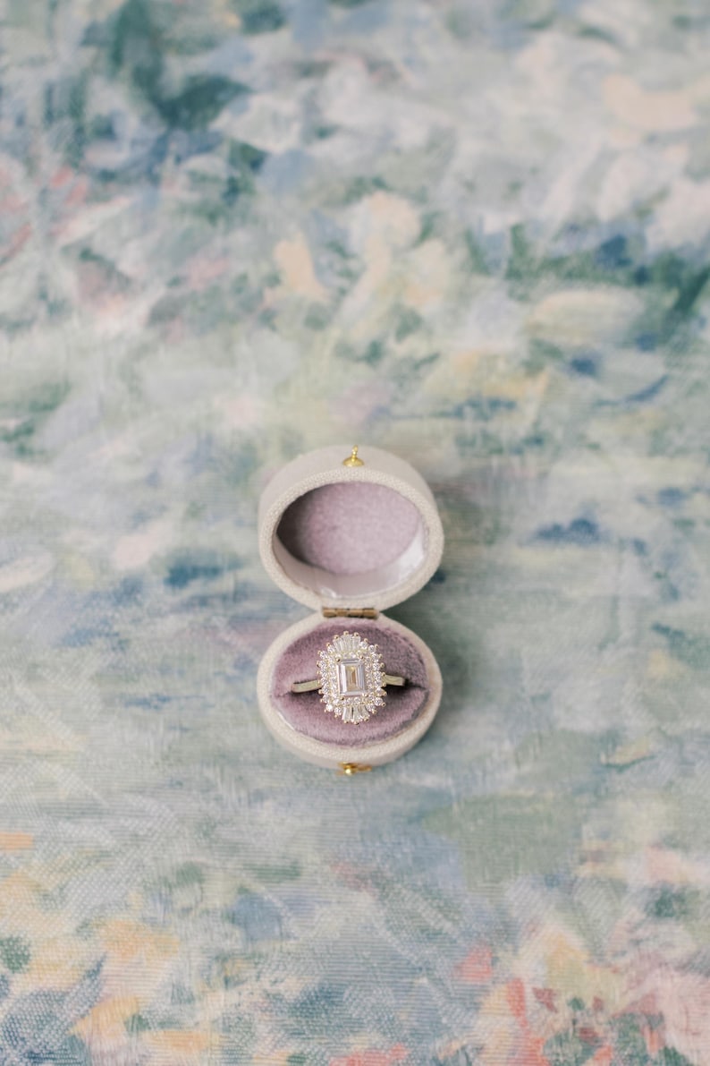 May include: A silver-toned ring featuring a central emerald-cut stone surrounded by a halo of smaller baguette and round-cut stones, resting inside a small, oval-shaped, light-colored velvet ring box with a soft, light purple interior lining.