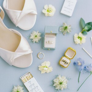 Puede incluir: Una vista cenital de detalles de boda sobre una superficie azul claro. Zapatos de sat&eacute;n blanco, anillos en cajas abiertas, flores y cajas de cerillas est&aacute;n dispuestos. La caja de cerillas dice "Beaufort, SC".