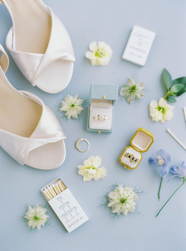 Puede incluir: Una vista cenital de detalles de boda sobre una superficie azul claro. Zapatos de sat&eacute;n blanco, anillos en cajas abiertas, flores y cajas de cerillas est&aacute;n dispuestos. La caja de cerillas dice "Beaufort, SC".