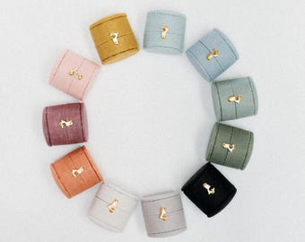May include: A collection of colorful ring boxes arranged in a circle. The boxes are in various pastel shades, including pink, yellow, and blue. Each box has a gold-colored clasp. The boxes are made of fabric and are likely used for jewelry storage.