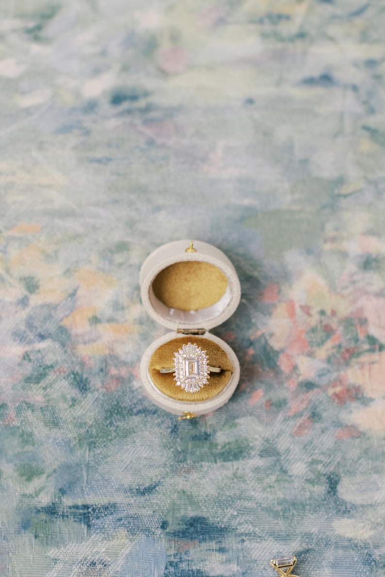 May include: A gold ring featuring a central emerald-cut diamond surrounded by a halo of smaller baguette and round diamonds, resting inside an oval-shaped, cream-colored velvet ring box with a mustard-yellow interior lining.