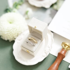 Puede incluir: Un juego de anillos de boda en una caja blanca, exhibido en un plato decorativo blanco. Un anillo es dorado, el otro plateado con peque&ntilde;as piedras. Un sello de cera con mango de madera est&aacute; a la derecha.