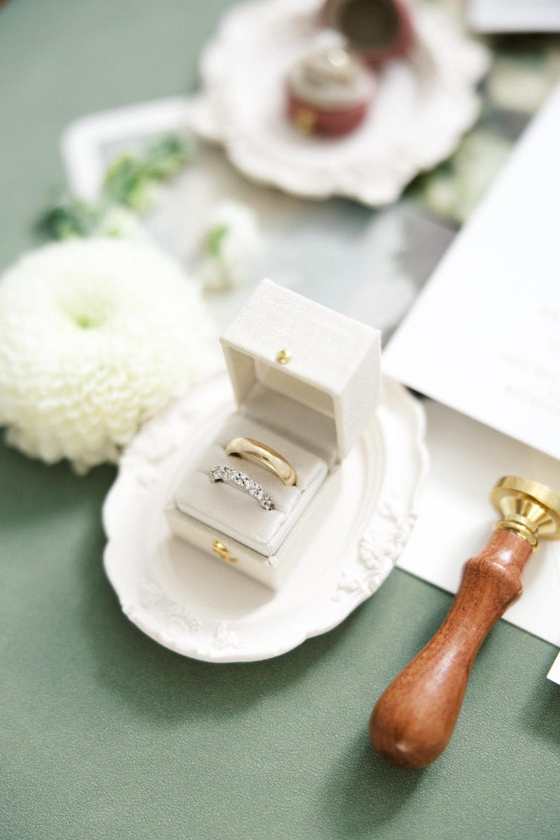 Puede incluir: Un juego de anillos de boda en una caja blanca, exhibido en un plato decorativo blanco. Un anillo es dorado, el otro plateado con peque&ntilde;as piedras. Un sello de cera con mango de madera est&aacute; a la derecha.