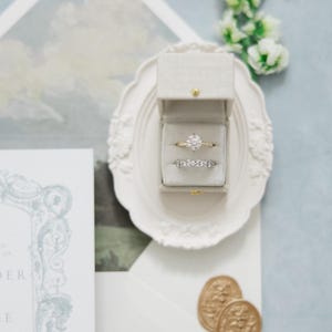 Puede incluir: Un juego de anillos de boda en una caja de terciopelo gris, exhibido en un plato decorativo blanco. El juego incluye una banda de oro con un diamante grande y redondo y una banda de eternidad de diamantes. Tambi&eacute;n se ven sellos de cera dorados y una invitaci&oacute;n de boda.