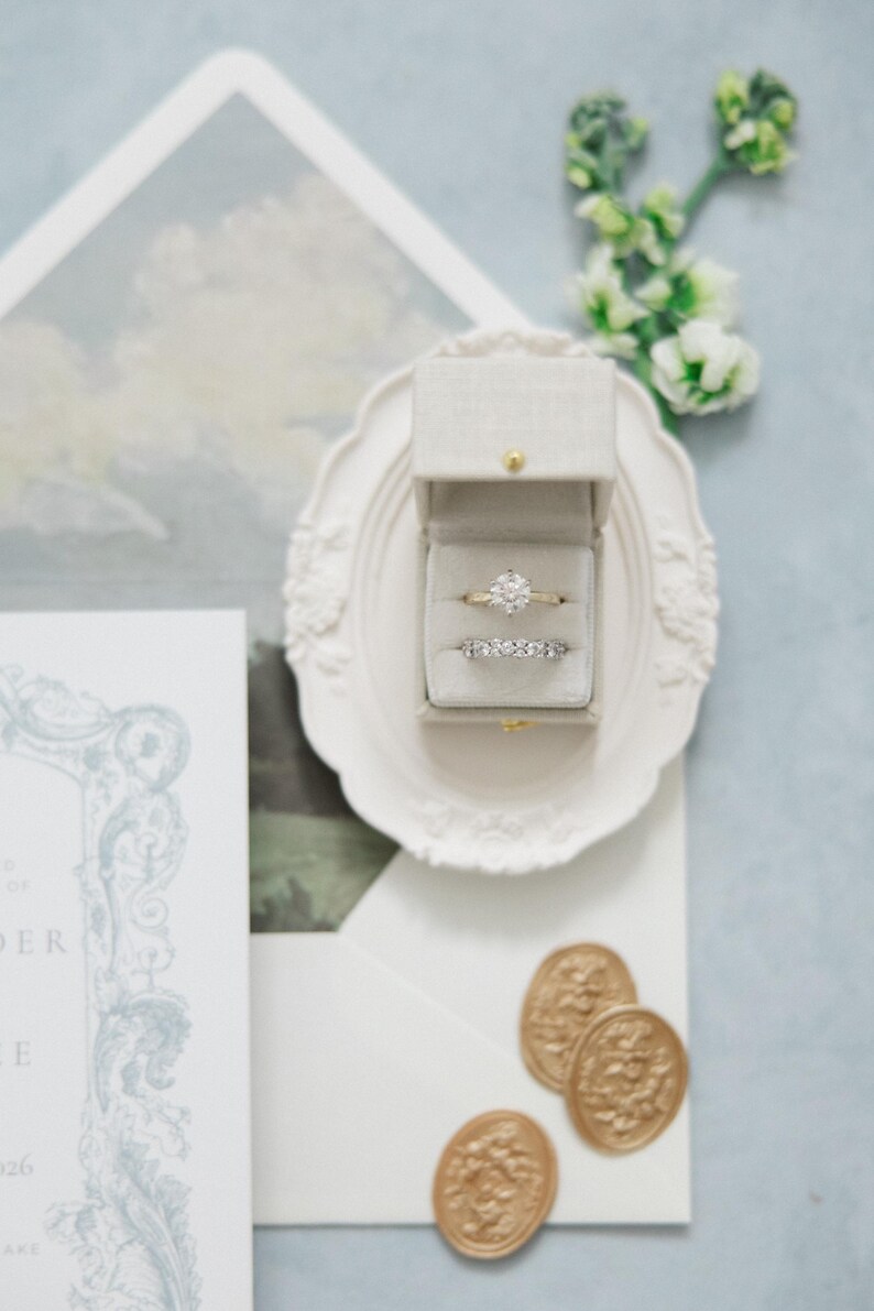 Puede incluir: Un juego de anillos de boda en una caja de terciopelo gris, exhibido en un plato decorativo blanco. El juego incluye una banda de oro con un diamante grande y redondo y una banda de eternidad de diamantes. Tambi&eacute;n se ven sellos de cera dorados y una invitaci&oacute;n de boda.