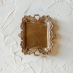 May include: A gold-toned decorative tray with an ornate, scrolled design. The tray has a rectangular shape and a smooth, slightly textured surface.
