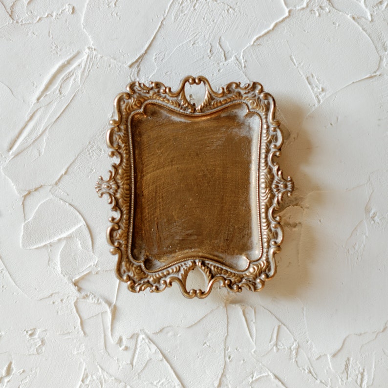 May include: A gold-toned decorative tray with an ornate, scrolled design. The tray has a rectangular shape and a smooth, slightly textured surface.