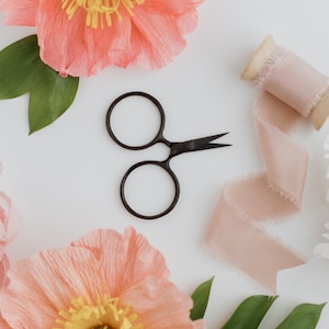 May include: Overhead shot of crafting supplies on a white surface. Includes black scissors, a spool of blush-colored ribbon, and coral and white paper flowers with green leaves. A visually appealing arrangement for creative projects.