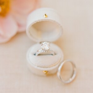May include: A round brilliant cut diamond engagement ring with a pav&eacute; band sits inside a white velvet ring box. A matching plain metal wedding band is positioned next to the open box. The jewelry features a polished silver-toned metal finish.