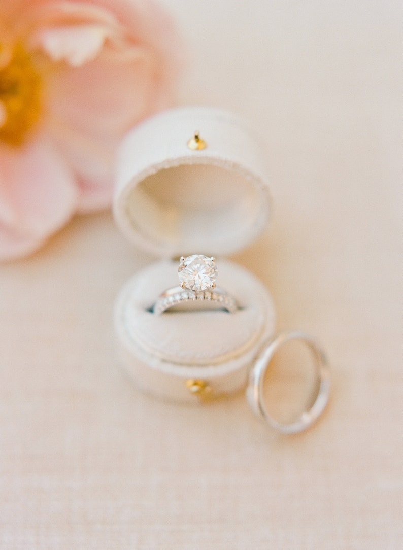 May include: A round brilliant cut diamond engagement ring with a pav&eacute; band sits inside a white velvet ring box. A matching plain metal wedding band is positioned next to the open box. The jewelry features a polished silver-toned metal finish.