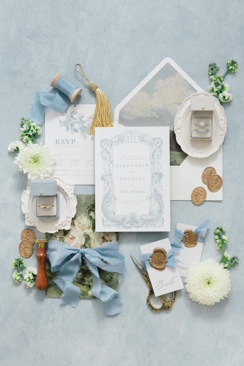 Puede incluir: Un elegante conjunto de papeler&iacute;a de boda con detalles en azul claro. Incluye tarjetas de invitaci&oacute;n, tarjetas de confirmaci&oacute;n de asistencia, sobres, anillos en cajas, sellos de cera dorados, cinta azul, tijeras y adornos florales. La invitaci&oacute;n dice "Alexander y Rosalee, 16 de mayo de 2016."