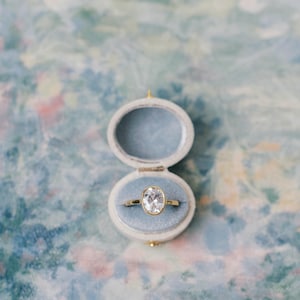 May include: A gold ring with an oval-cut clear gemstone set in a bezel, resting inside a small, oval-shaped, light blue velvet-lined ring box. The ring features a polished gold band and a single, prominent, faceted white stone at its center.