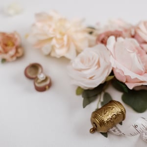 May include: A romantic still life featuring a velvet ring box with a diamond ring, blush-colored roses, and a vintage gold bell. A measuring tape and scattered flower petals complete the scene, evoking a sense of elegance and celebration.